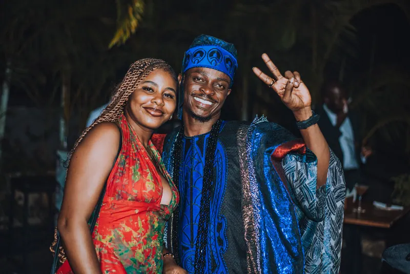 A woman in a floral dress and a man in a blue agbada lean together, him throwing a peace sign