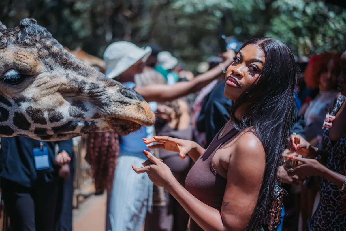 A woman in a brown top glances at the camera while a giraffe leans in to be fed