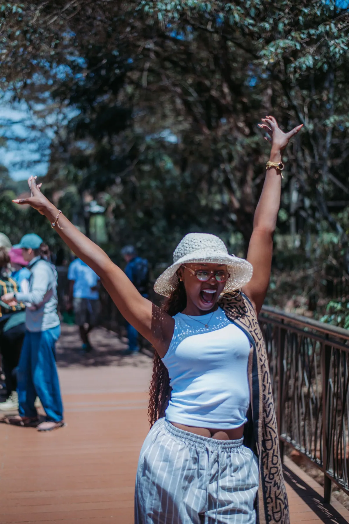A woman in a crochet hat throws both arms in the air on the Giraffe Centre boardwalk