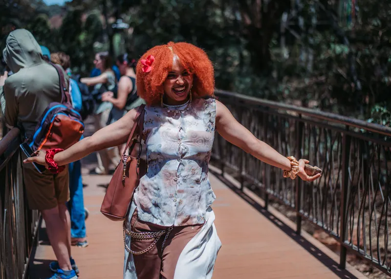 A woman with a red afro and a flower in her hair spreads her arms wide, laughing, on the boardwalk