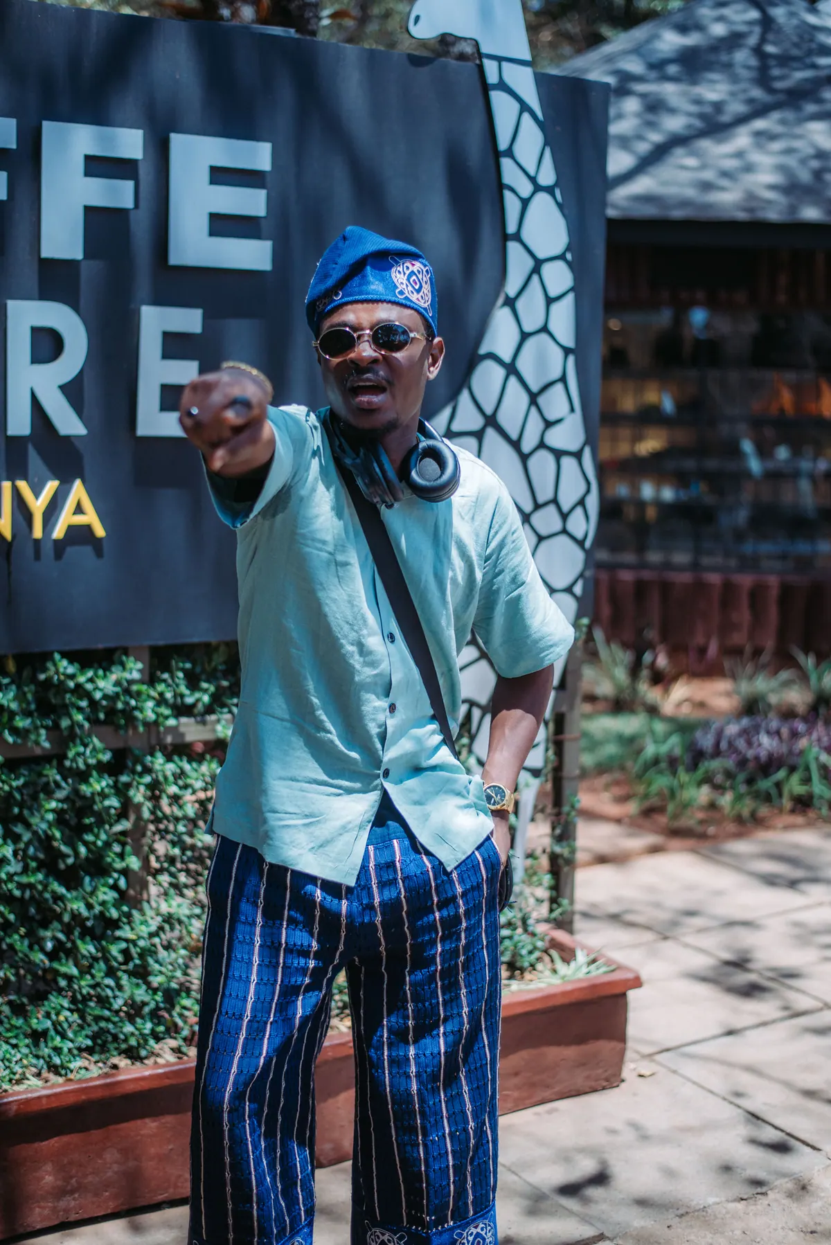 A man in a blue embroidered cap and striped trousers points at the camera in front of the Giraffe Centre sign
