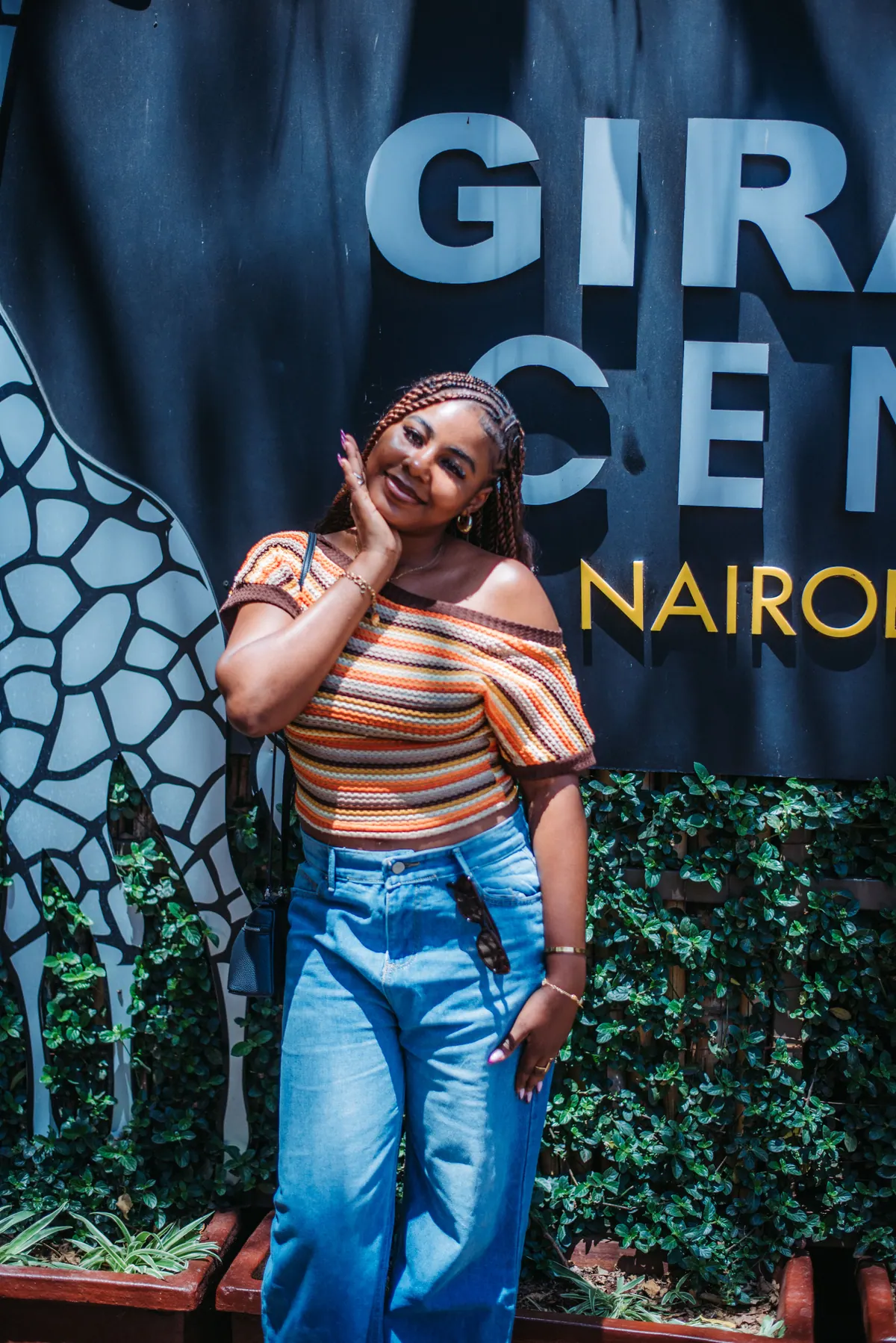 A woman in a striped top and wide jeans poses with a hand to her hair in front of the Giraffe Centre sign