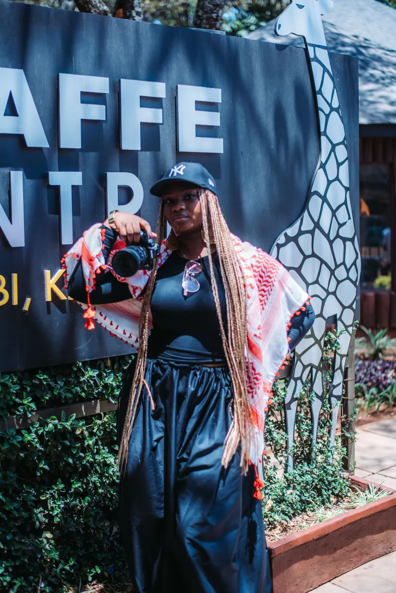 A woman in a navy cap with a pink patterned scarf over her shoulders stands by the Giraffe Centre sign