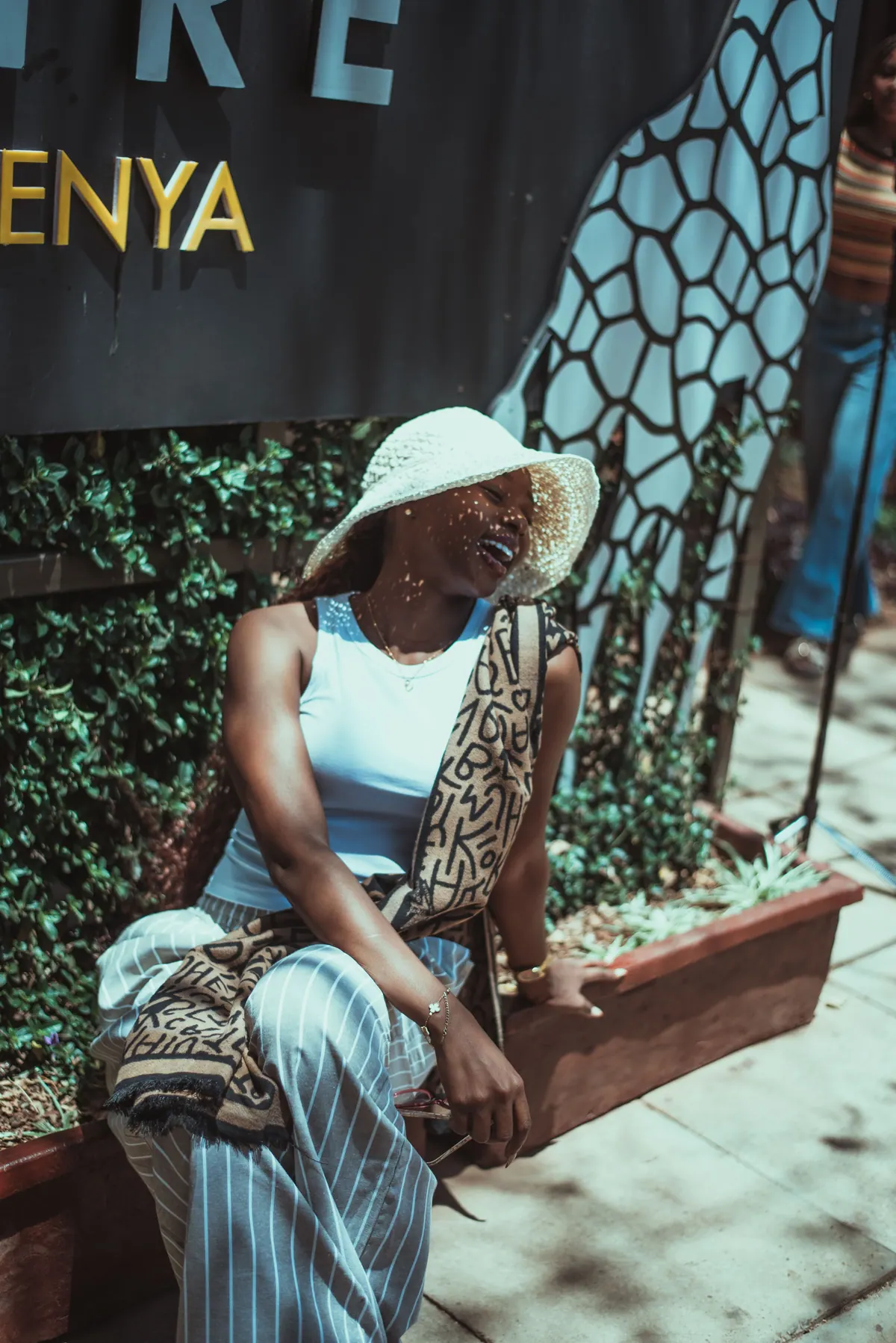 A woman in a crochet hat laughs with her eyes closed, sitting under the Giraffe Centre sign