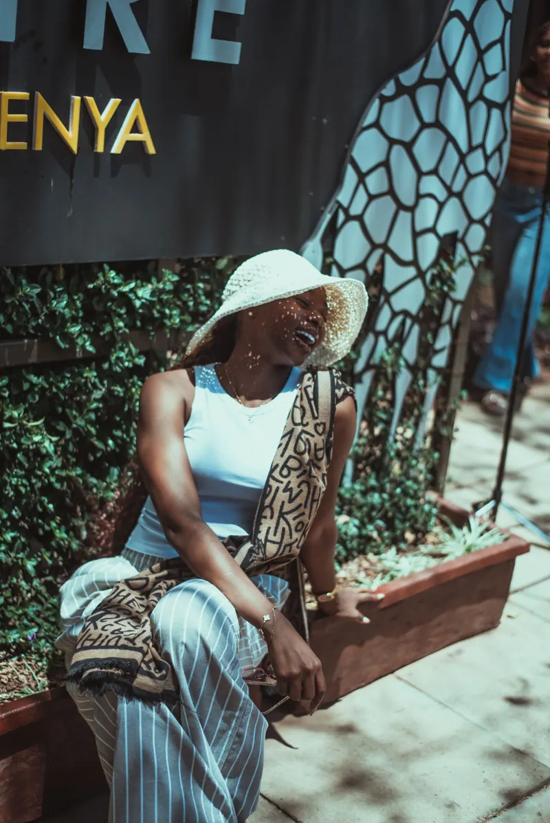A woman in a crochet hat laughs with her eyes closed, sitting under the Giraffe Centre sign