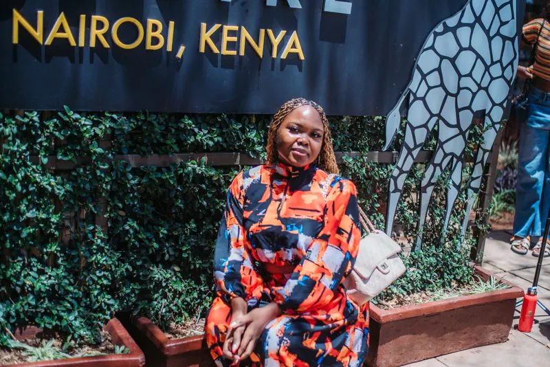 A woman in an orange and blue print set sits under the Giraffe Centre sign