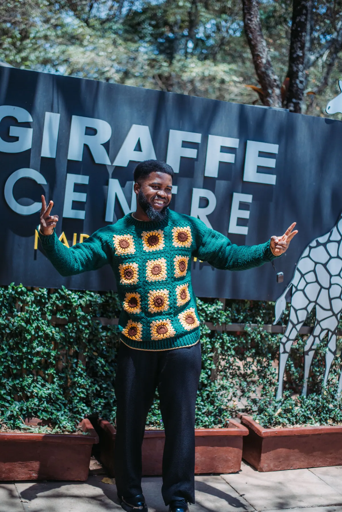 A man in a green crochet cardigan spreads his arms in front of the Giraffe Centre sign