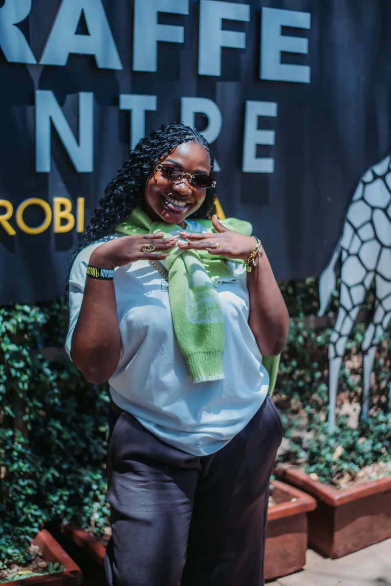 A woman in tinted glasses with a green jumper over her shoulders smiles in front of the Giraffe Centre sign