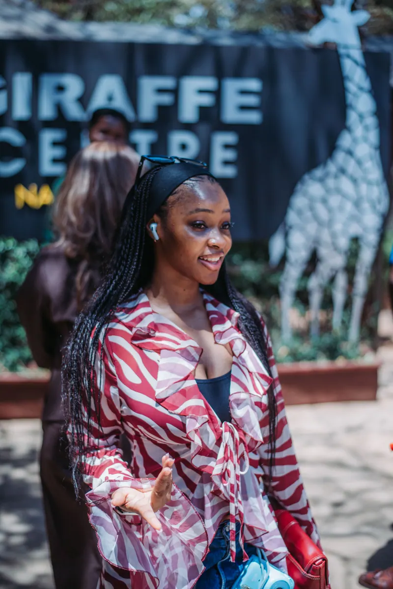 A woman with long braids in a red and white zebra-print shirt smiles outside the Giraffe Centre