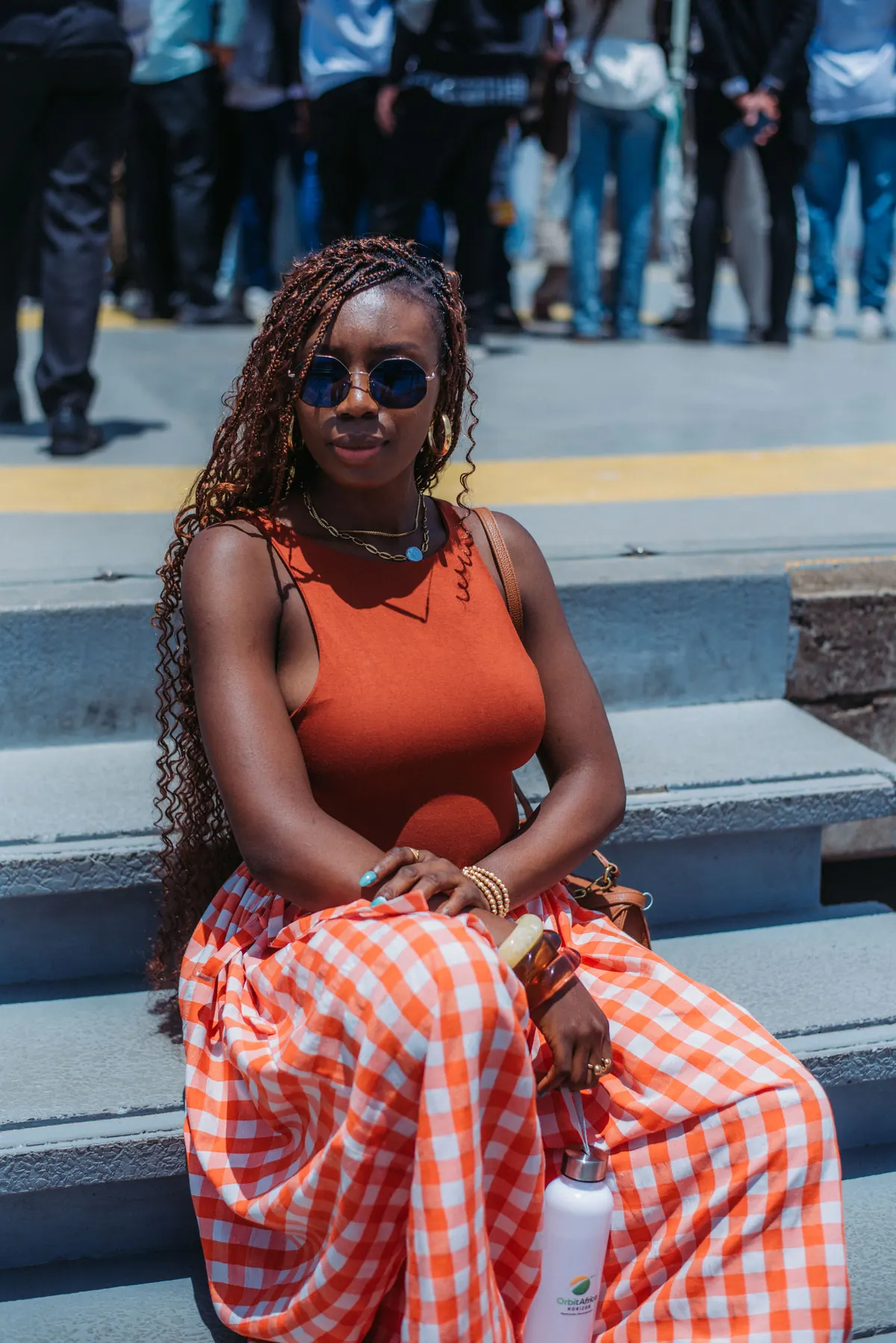 A woman in round sunglasses, a rust top and an orange gingham skirt sits on steps holding an Orbit Africa Horizon water bottle