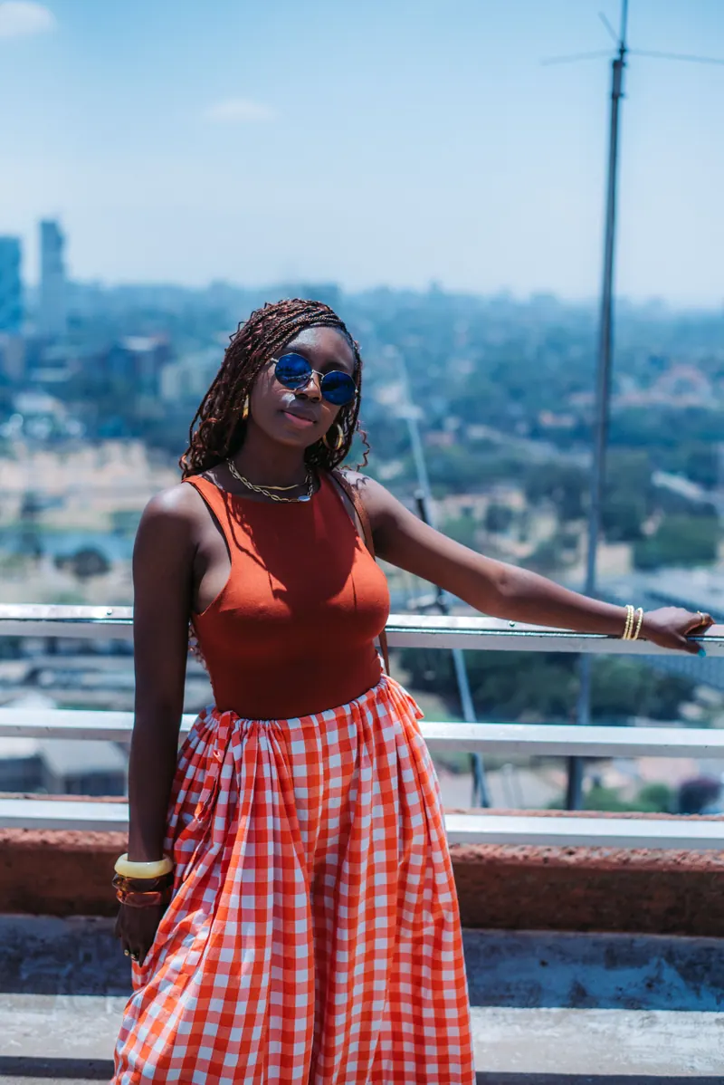 A woman in an orange gingham skirt leans on the rooftop railing at KICC with Nairobi spread out below