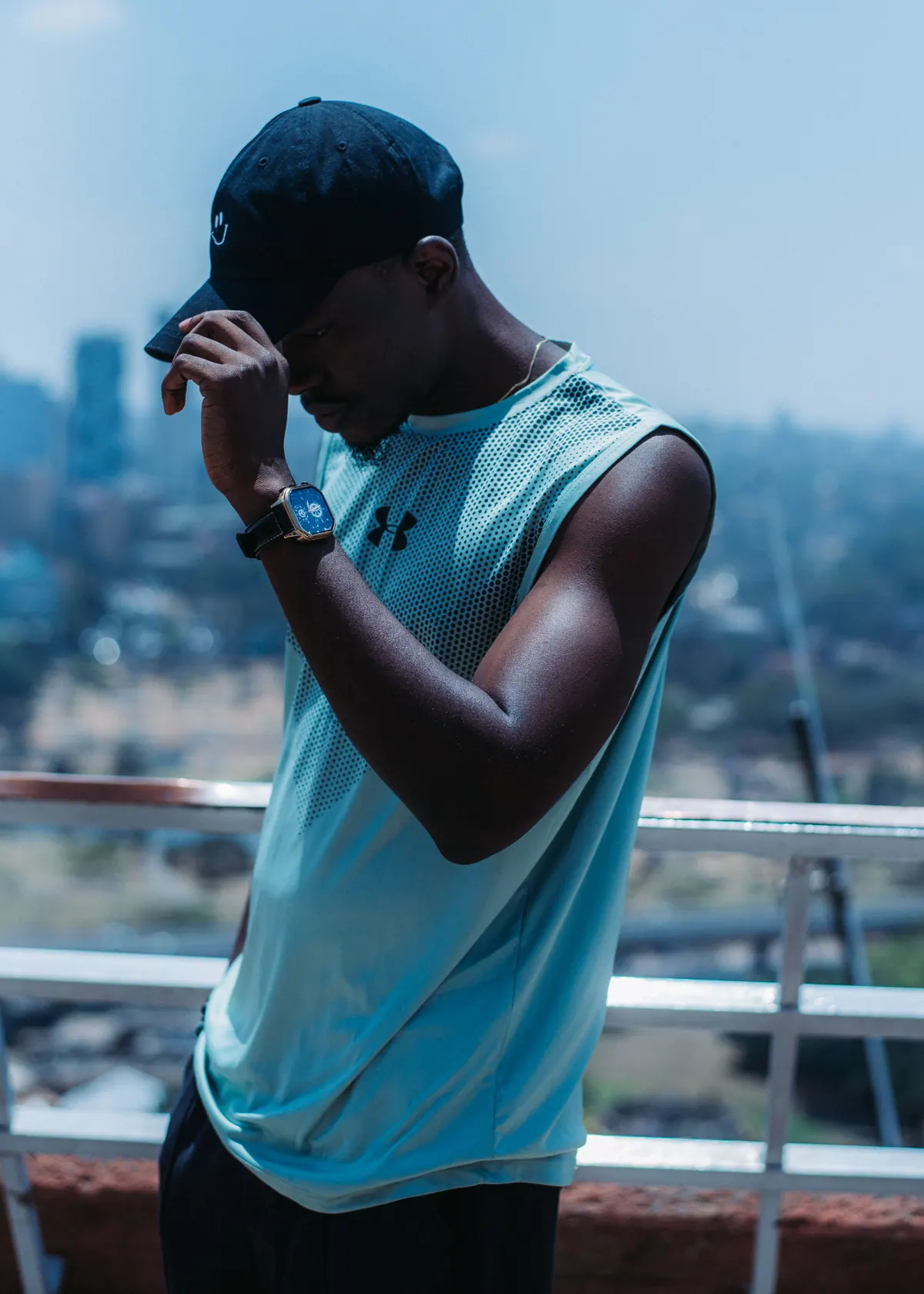 A man in a mint sleeveless top tips his black cap down on the KICC rooftop, the Nairobi skyline behind him
