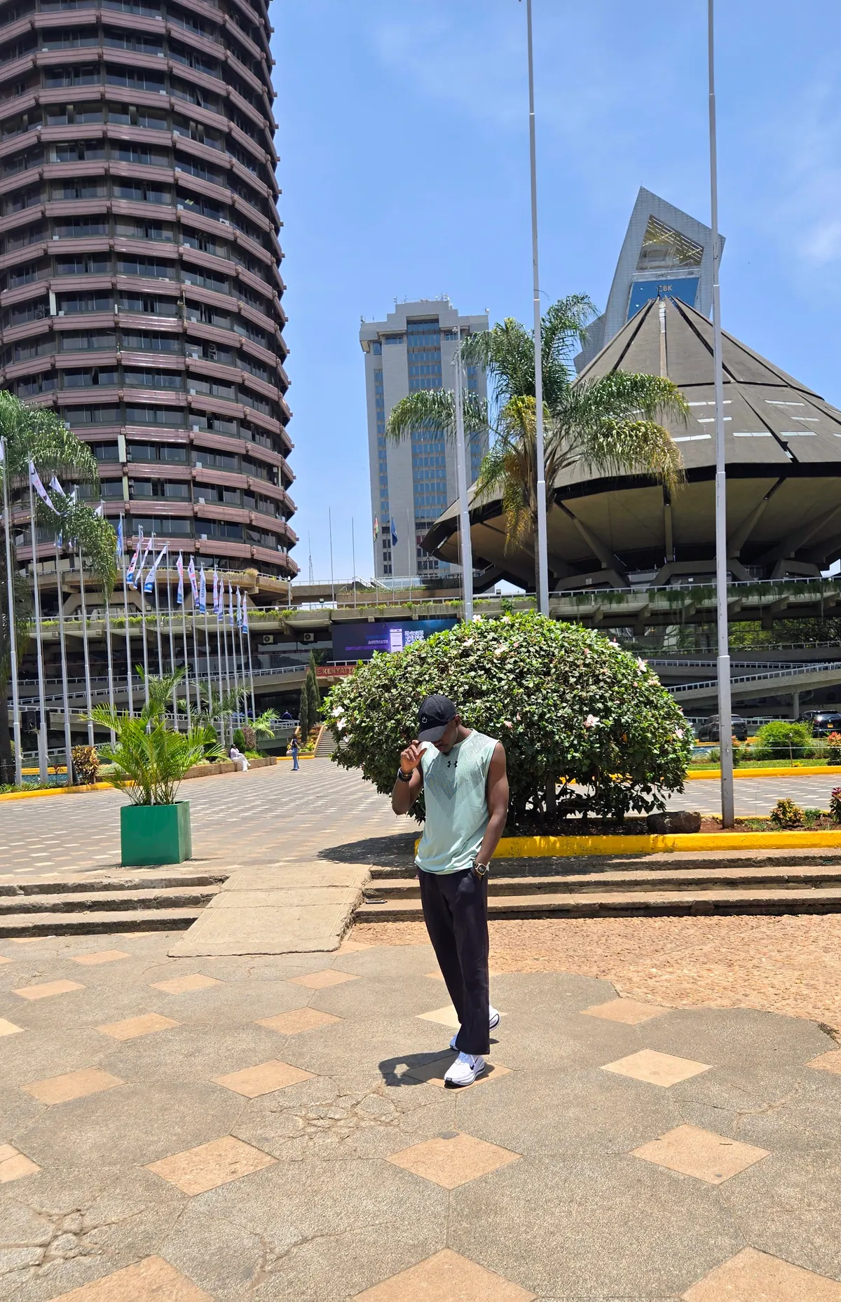 A man in a mint sleeveless top tips his cap in the KICC forecourt, the tower and amphitheatre behind him