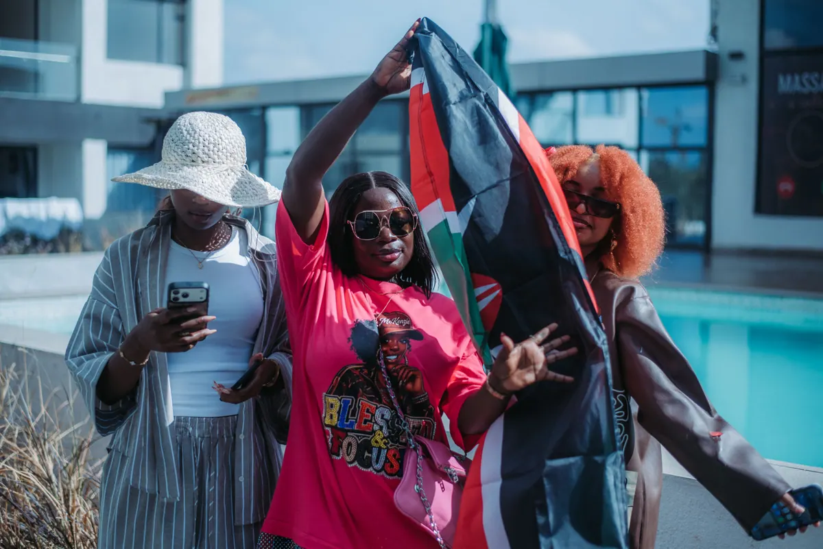 Three women pose by the pool, one lifting a Kenyan flag overhead