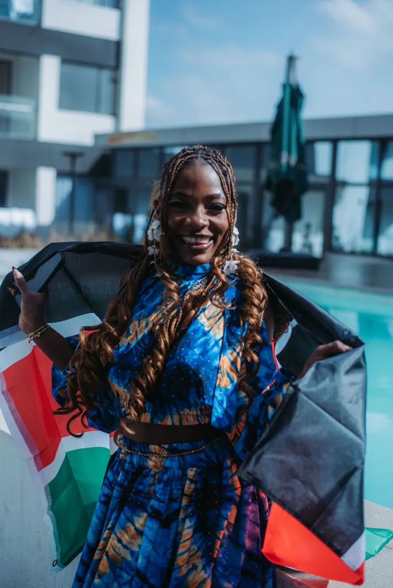 A woman in a blue and orange print two-piece smiles with a Kenyan flag draped behind her shoulders by the pool
