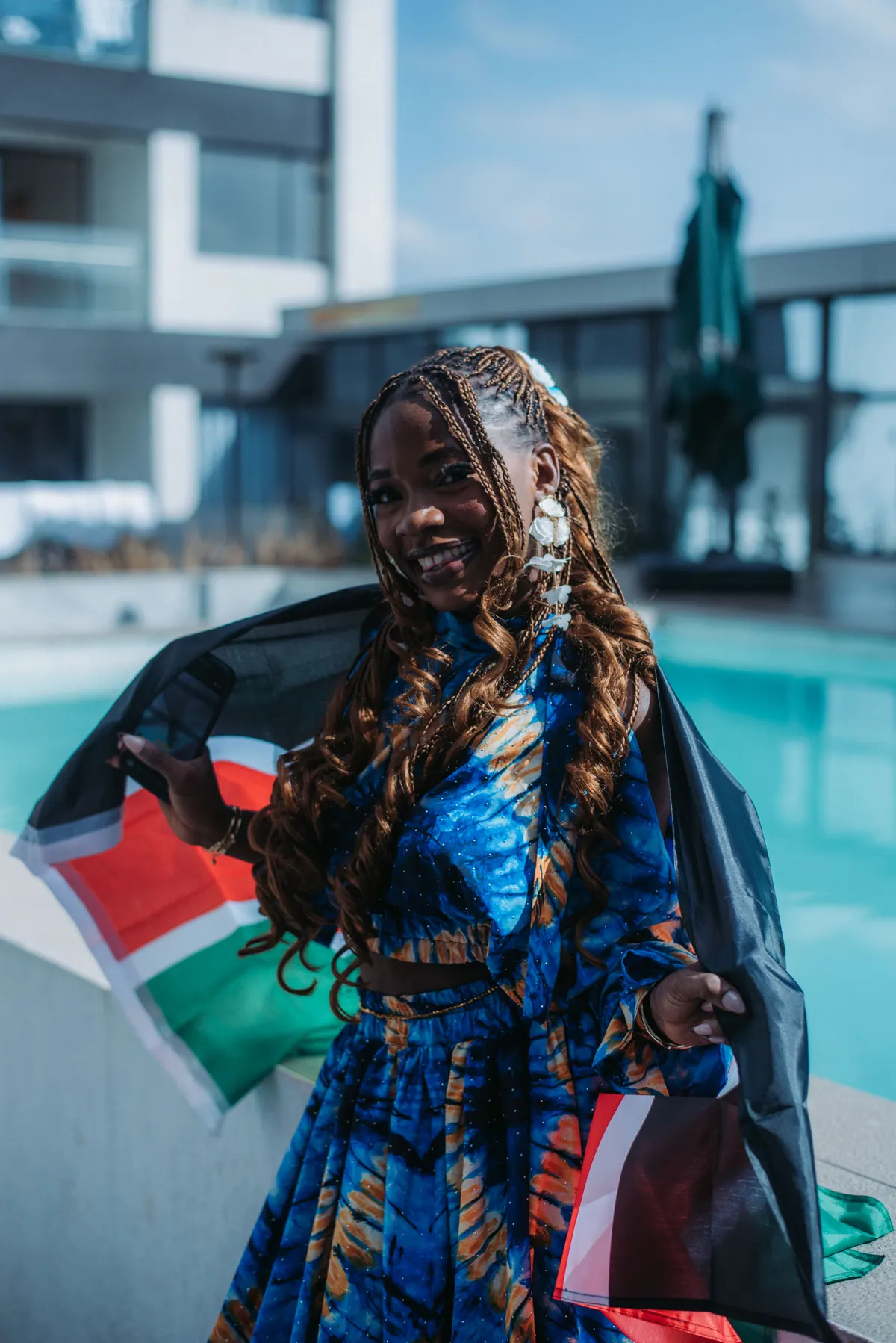 A woman with long curled braids and flower earrings wraps a Kenyan flag around herself beside the pool