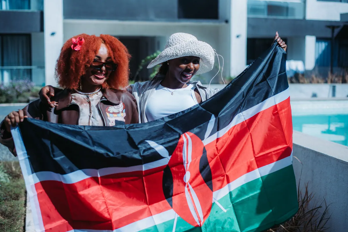Two women hold a Kenyan flag out between them beside the pool, one with a red afro, one in a crochet hat