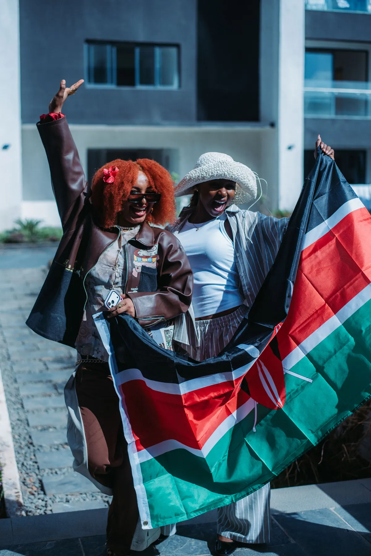 A woman with a red afro raises an arm in the air while her friend holds a Kenyan flag beside her