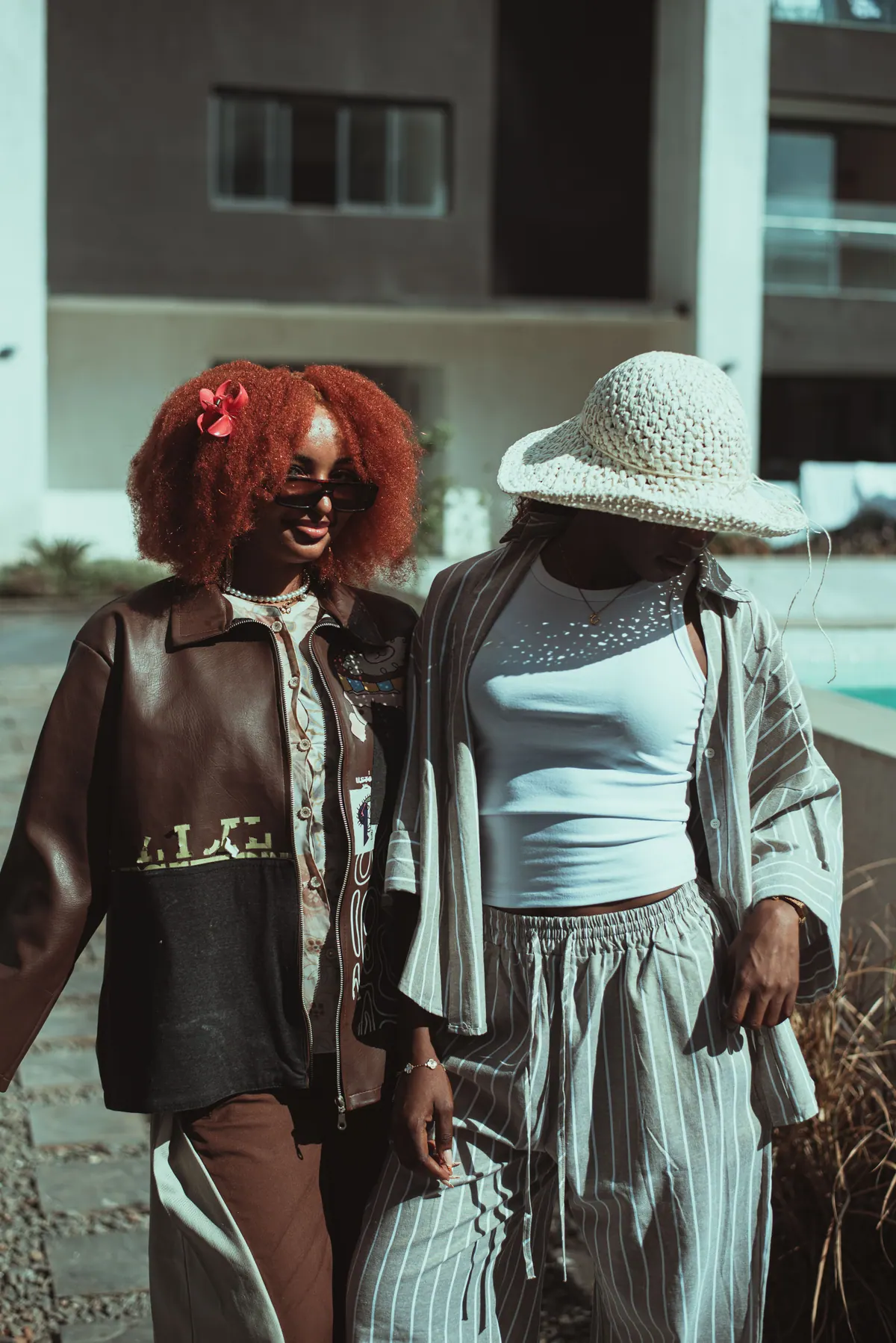 Two women walk arm in arm past the pool, one with a red afro and a brown leather jacket, the other in a crochet sun hat and striped linen