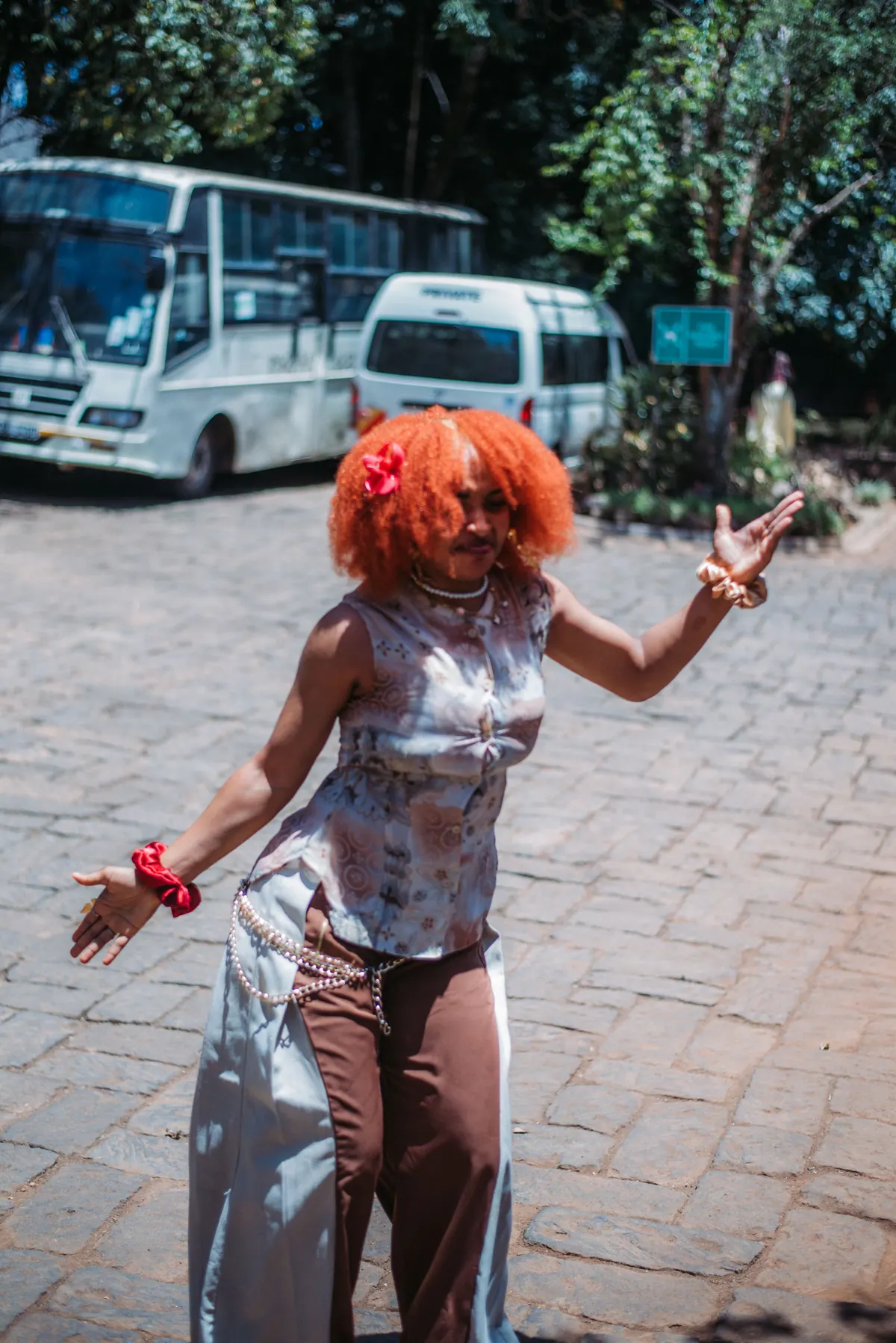 A woman with a red afro dances across a car park with her arms out