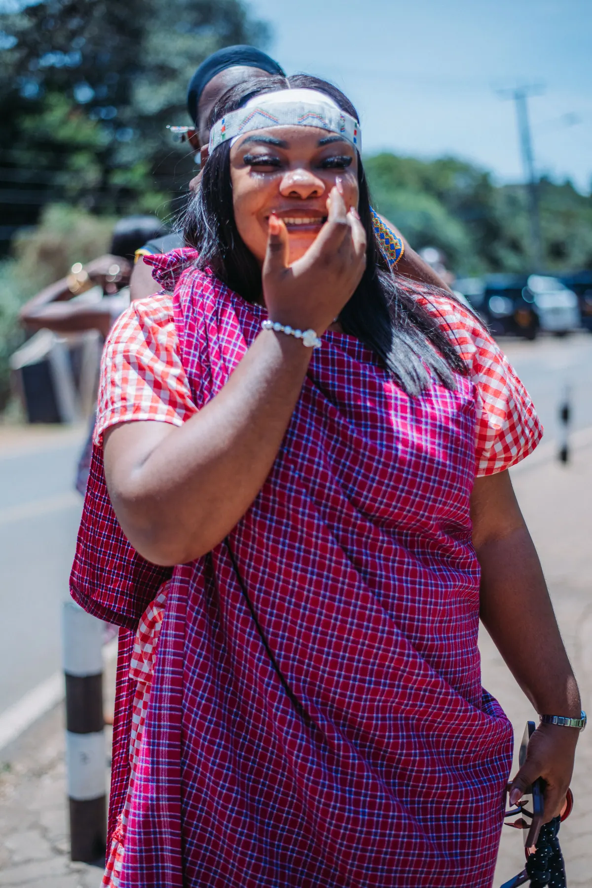 A woman in a purple checked shuka and beaded headband laughs behind her hand at the roadside