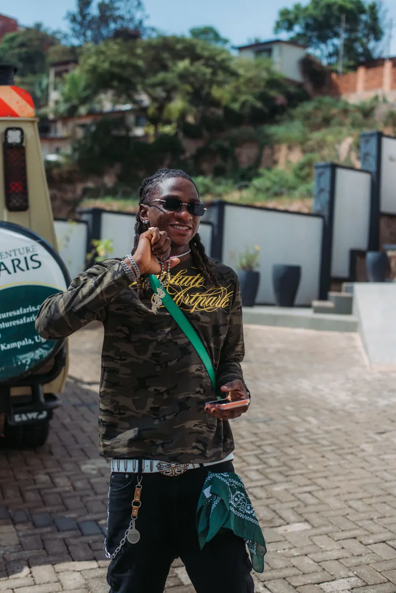 A man with locs, dark glasses and a camouflage top grins beside a safari vehicle on a paved forecourt