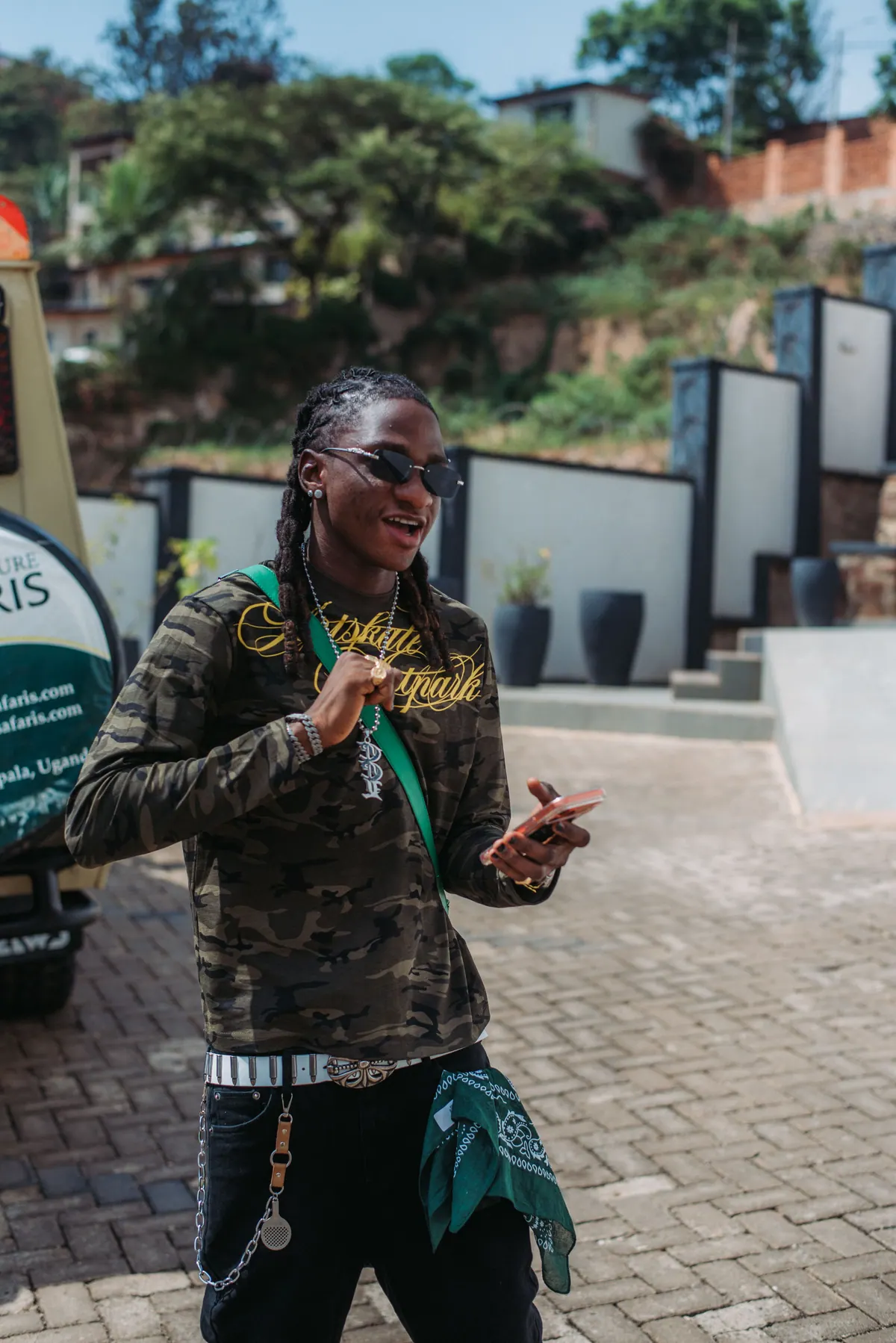 A man in a camouflage top and green bandana laughs while holding his phone on the forecourt