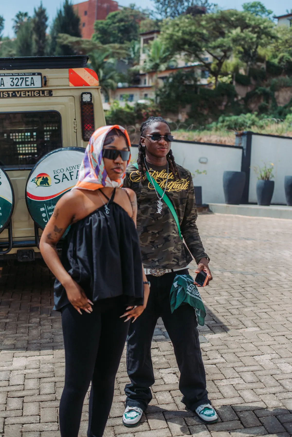A woman in a headscarf and a man in a camouflage top stand close together in front of a safari vehicle