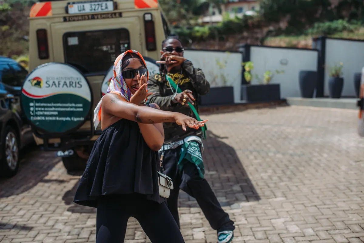 A woman in a patterned headscarf and visor sunglasses dances in front of a safari vehicle while a man strikes a pose behind her