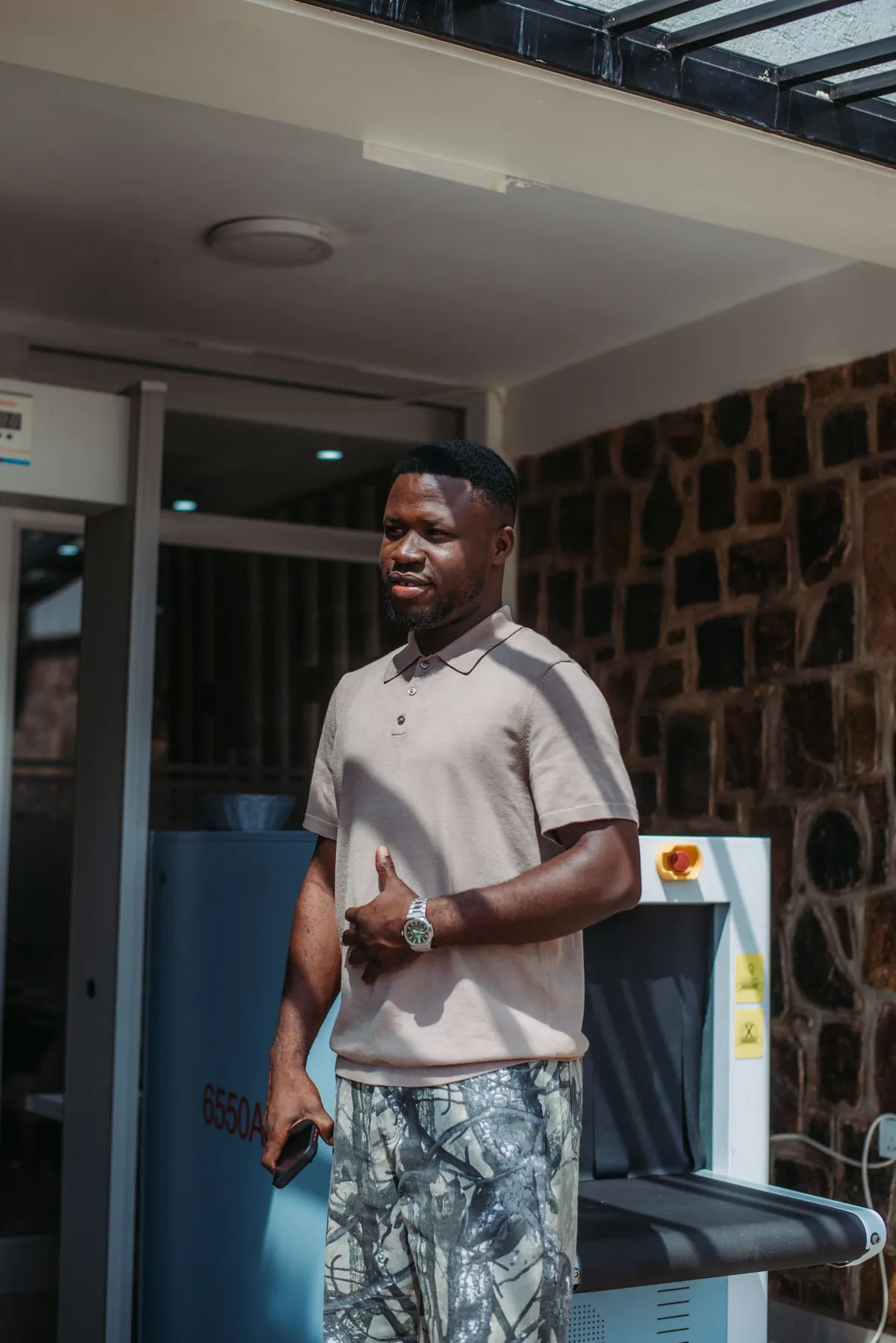 A man in a beige polo and camouflage shorts stands in the sun outside a hotel entrance