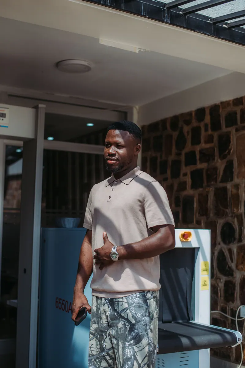 A man in a beige polo and camouflage shorts stands in the sun outside a hotel entrance