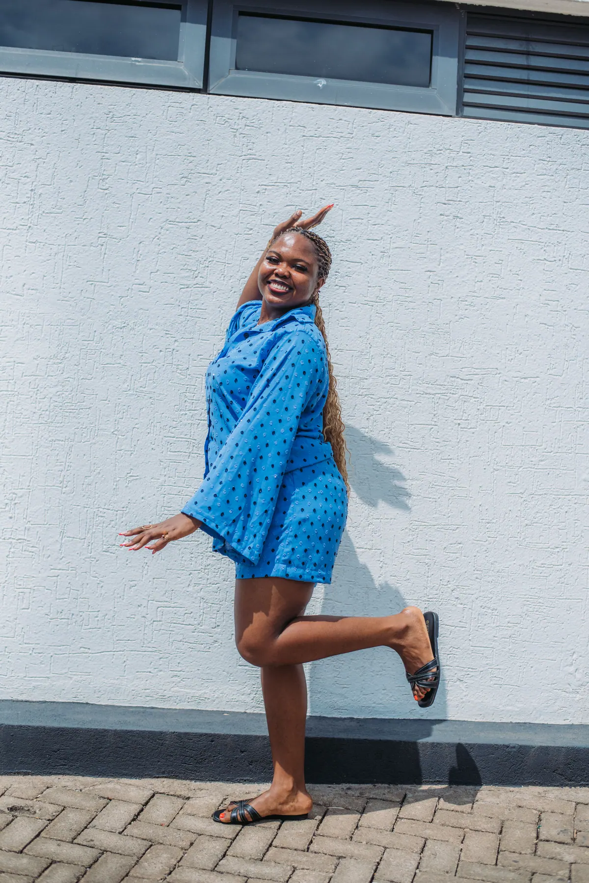 A woman in a bright blue shirt and shorts set kicks one leg up against a white wall