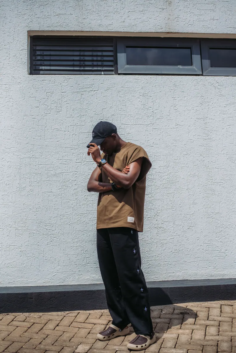 A man in a brown T-shirt and black cap looks down at his watch against a white wall