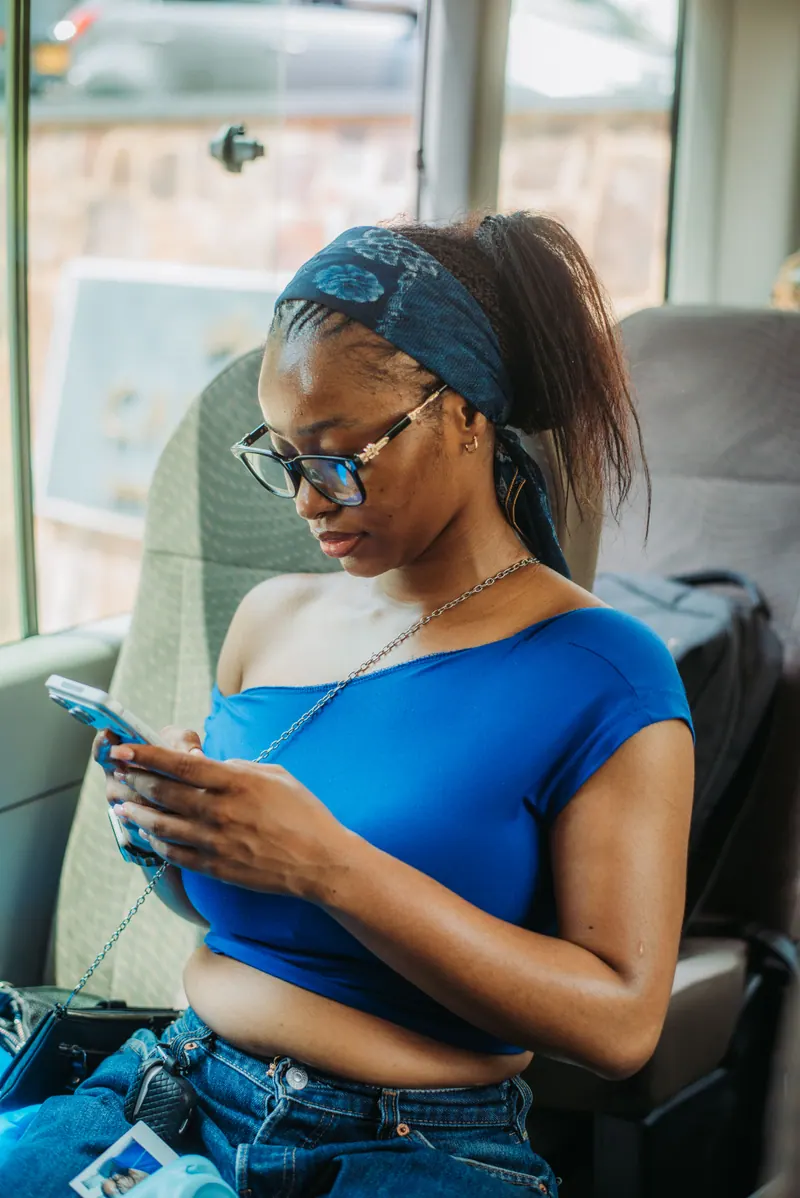 A woman in glasses, a blue headband and a blue top checks her phone in a window seat on the bus