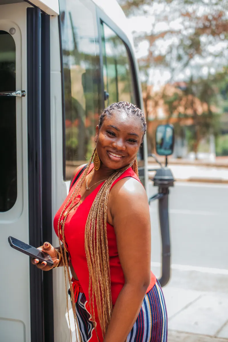A woman with long braids in a red top smiles as she steps off the bus
