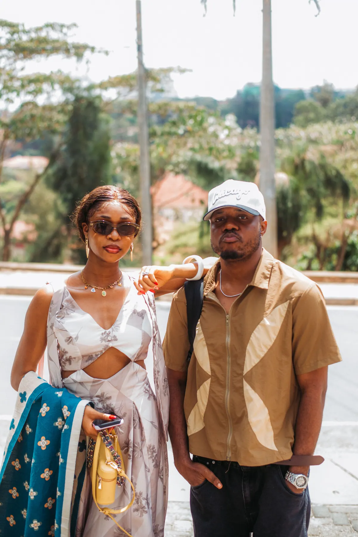 A woman in a white print set and a man in a white cap and tan shirt stand together by the roadside