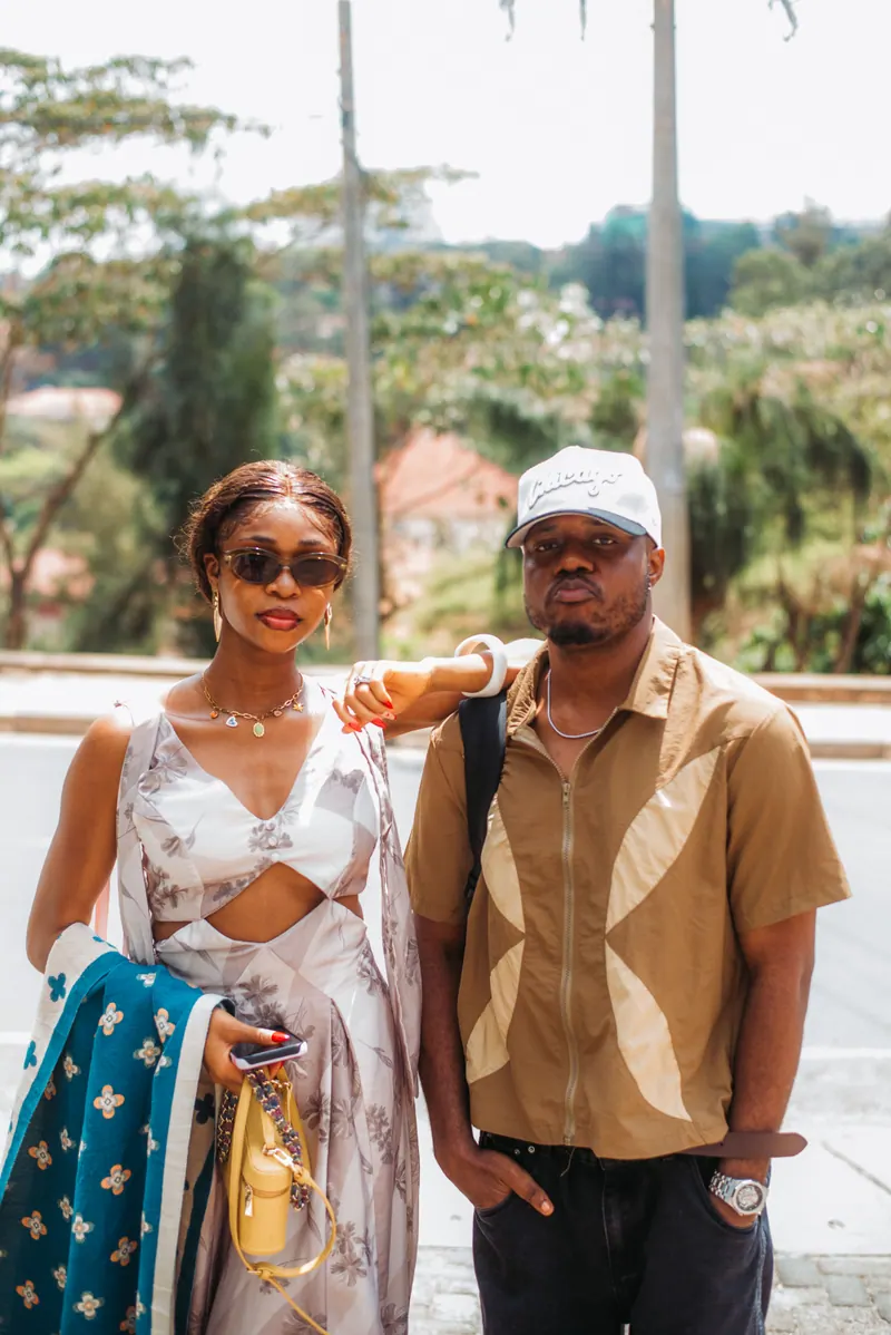 A woman in a white print set and a man in a white cap and tan shirt stand together by the roadside