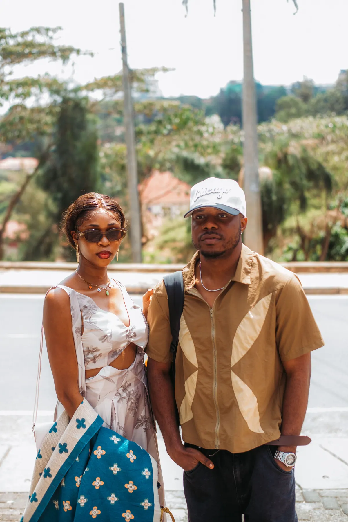 A woman in a white print set and a man in a white cap and tan shirt pose together at the roadside