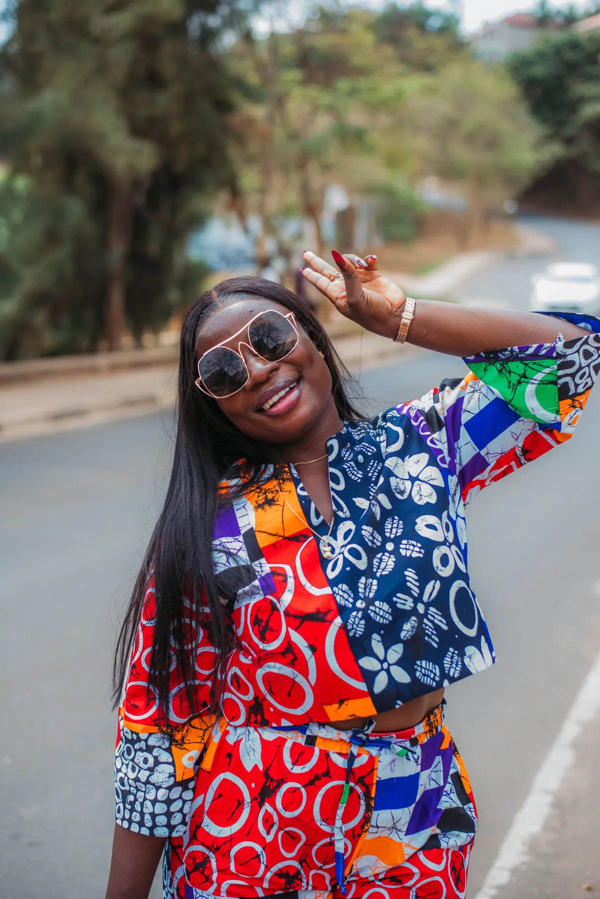 A woman in sunglasses and a red and blue patchwork print dress throws a peace sign on a quiet road