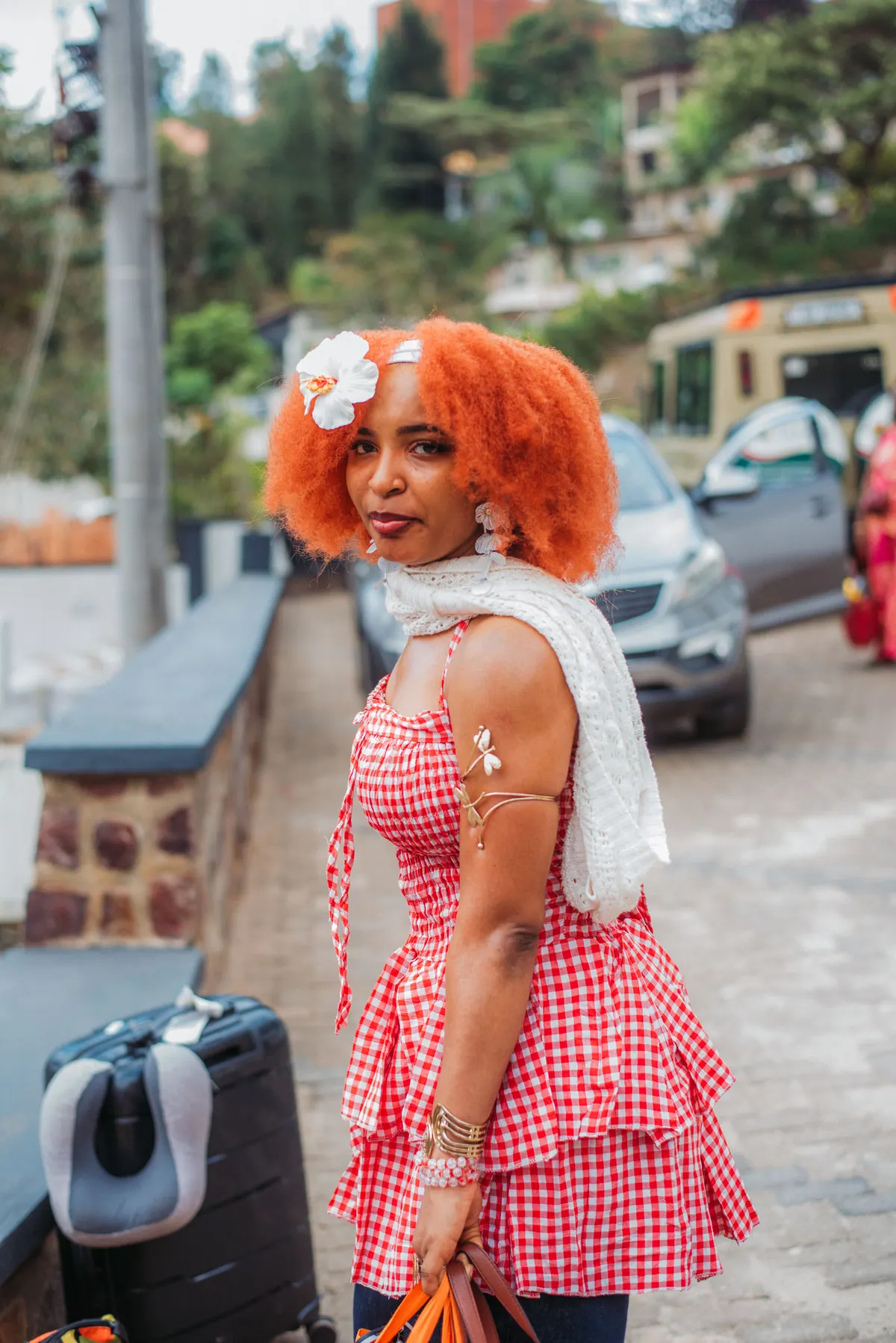 A woman with a red afro, a flower in her hair and a pink gingham dress stands by her suitcase