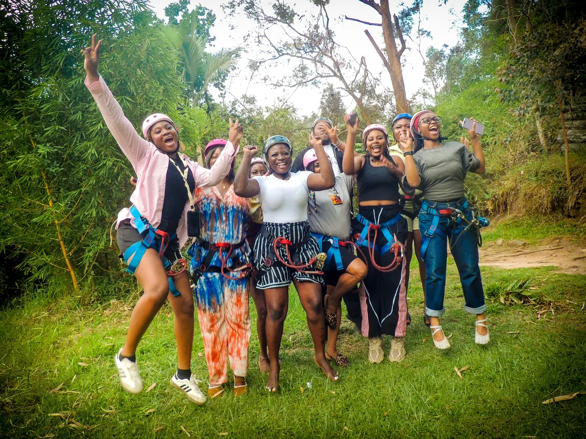 The zip-line crew in harnesses and pink helmets jump and cheer on the grass at Lake Mutanda