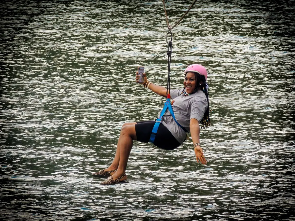 A woman in a pink helmet takes a selfie mid-air on the zip line, skimming the lake with her shoes