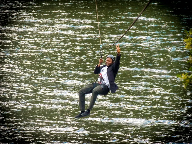 A man in a jacket throws a peace sign as he rides the zip line over the lake