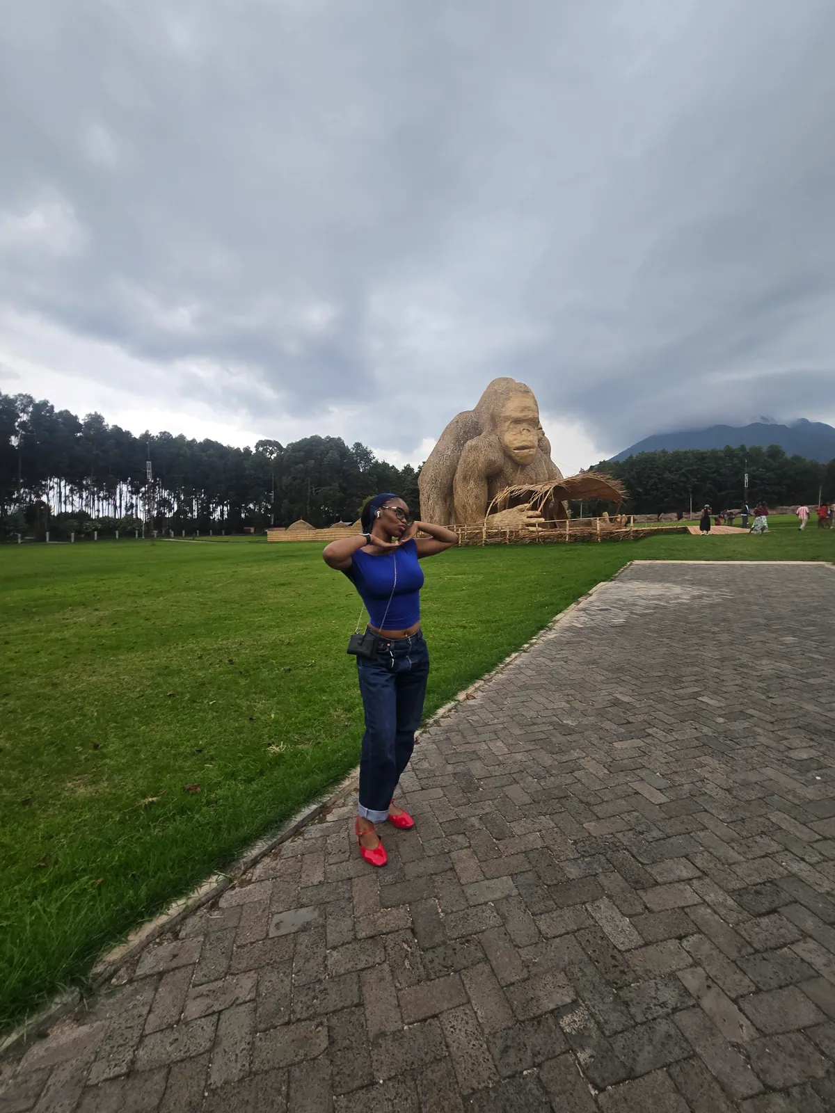 A woman in a blue top and jeans poses in front of a giant woven bamboo gorilla, with a volcano under cloud behind