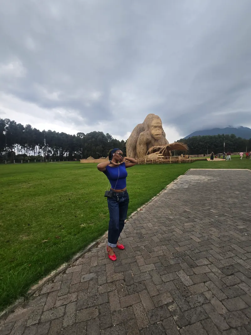 A woman in a blue top and jeans poses in front of a giant woven bamboo gorilla, with a volcano under cloud behind