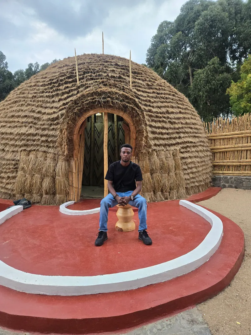 A man in a black T-shirt and jeans sits on a carved stool outside a thatched Rwandan house